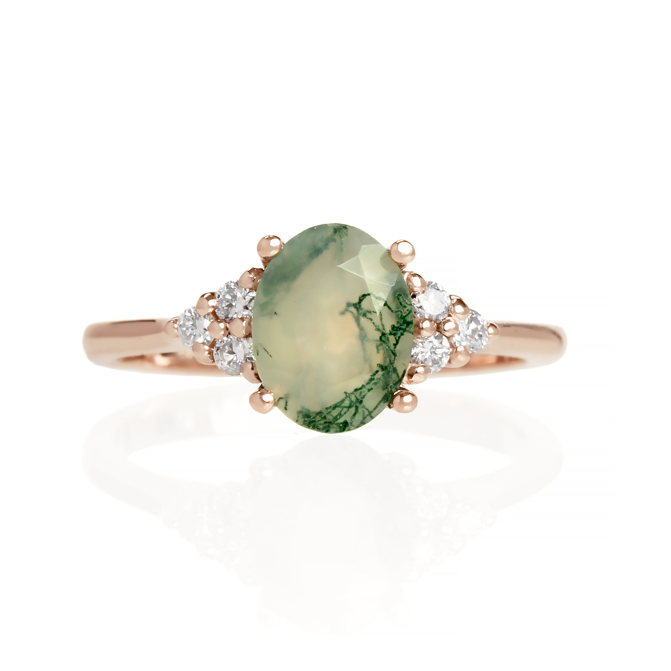 A product photo of a rose gold moss agate and diamond trio ring sitting on a white background. The oval-cut, naturally-included moss agate gemstone stands in stark contrast to the clusters of three classic white diamond stones on either side.