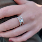 A product picture of a brilliant white moissanite engagement ring sitting on a woman's finger. The central moissanite jewel reflects several vibrant colours off of its many angles, while white jewelled detailing hugs the rims of the split 9k white-gold band.