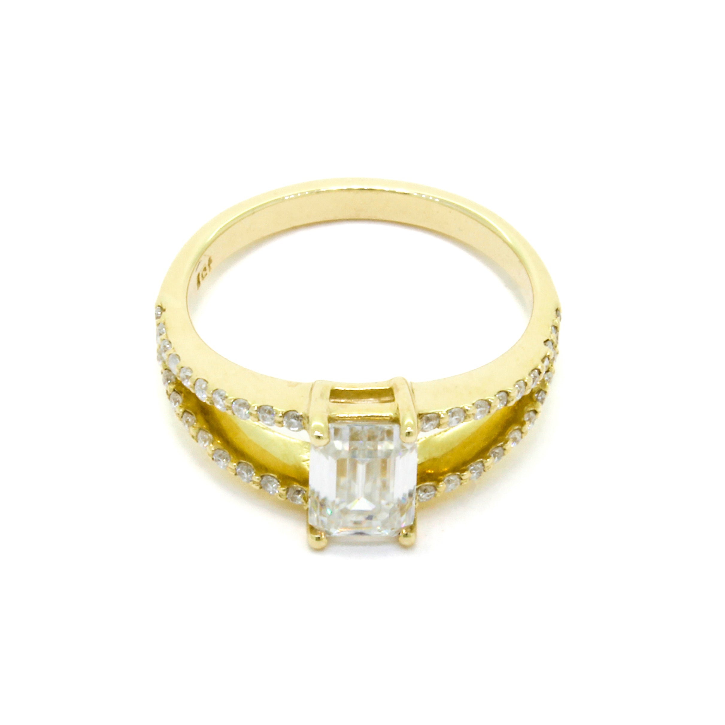 A product photo of a moissanite ring in 9k yellow gold sitting on a clear white background. The reflective emerald-cut moissanite jewel sits in the centre. A simple yellow gold band becomes embedded with diamonds the closer it gets towards the jewel, and splits into two bands on either side, both meeting in the centre of the ring to hold the moissanite in place.