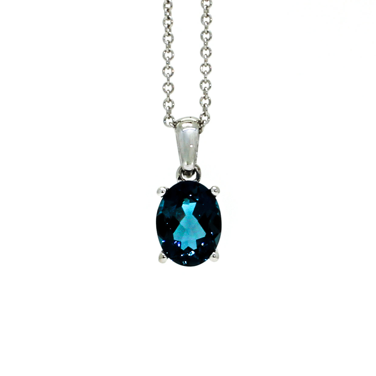 A product photo of a white gold london blue topaz pendant sitting against a white background. The impressively large and deeply-coloured oval-cut stone is contrasted by its overall minimalistic design, 4 simple white gold claws hold the gem in place. It is suspended by a simple white gold chain.