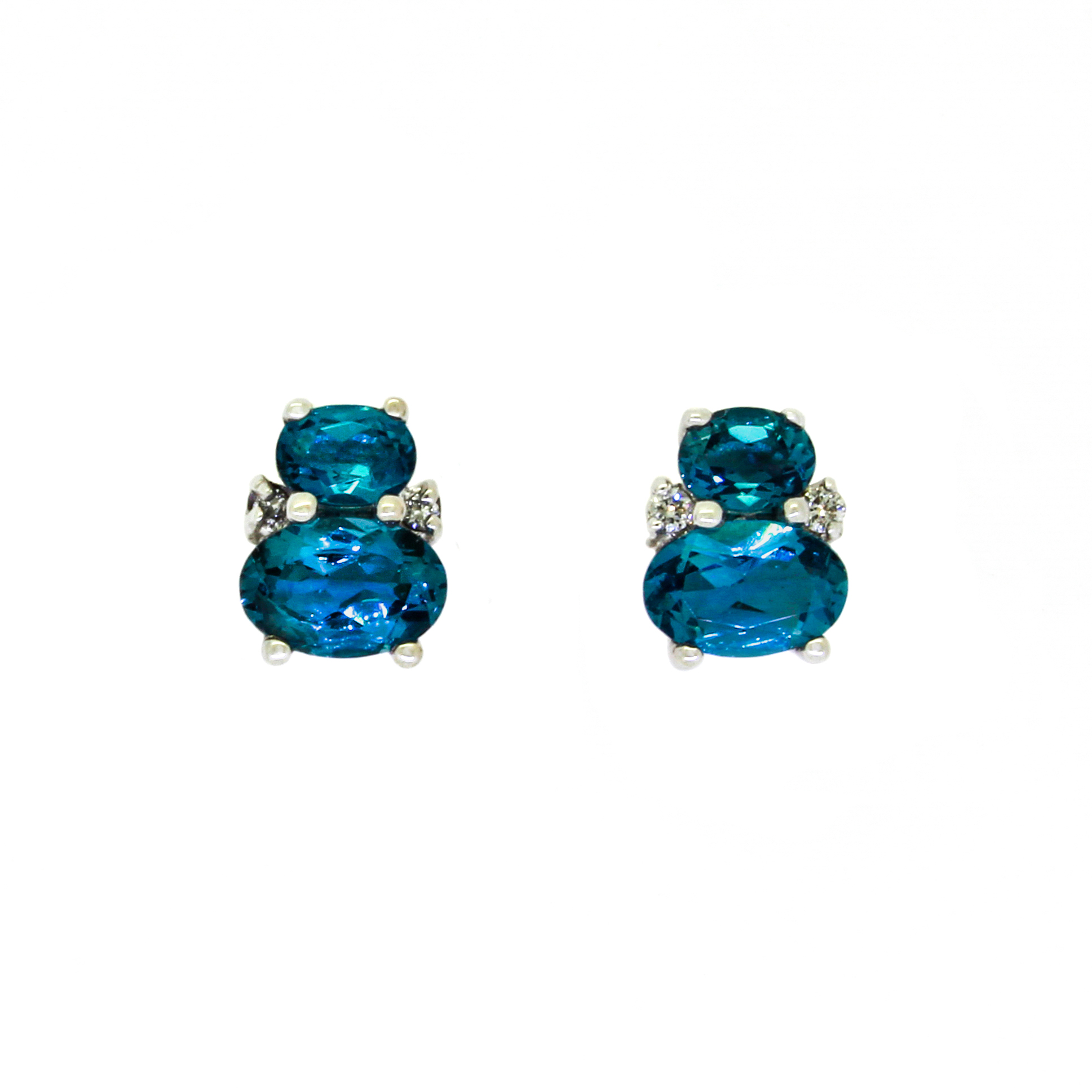 A product photo of a pair of white gold london blue topaz earrings sitting against a white background. One smaller oval is stacked upon a larger oval on each earring, with the area where the two stones make contact decorated by a single small diamond on either side.