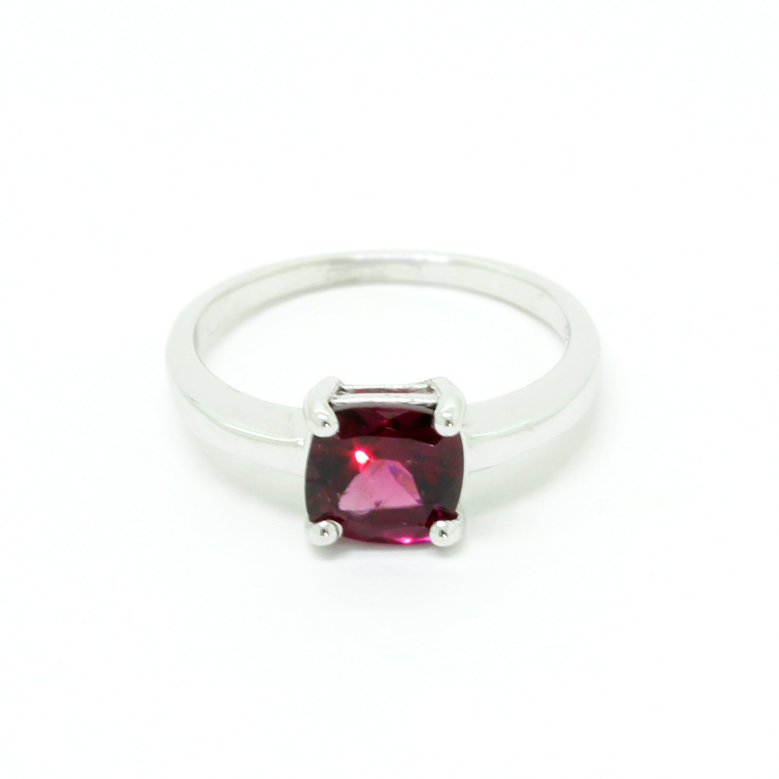 A product photo of a silver ring with a cushion-cut rhodalite centre stone sitting on a white background. The silver band is simple and smooth, connecting on either side of a square cushion-cut rhodalite stone held in place by four delicate silver claws. The rhodalite jewel is a deep velvet purple colour, reflecting a warm plum colours across its multi-faceted surface.