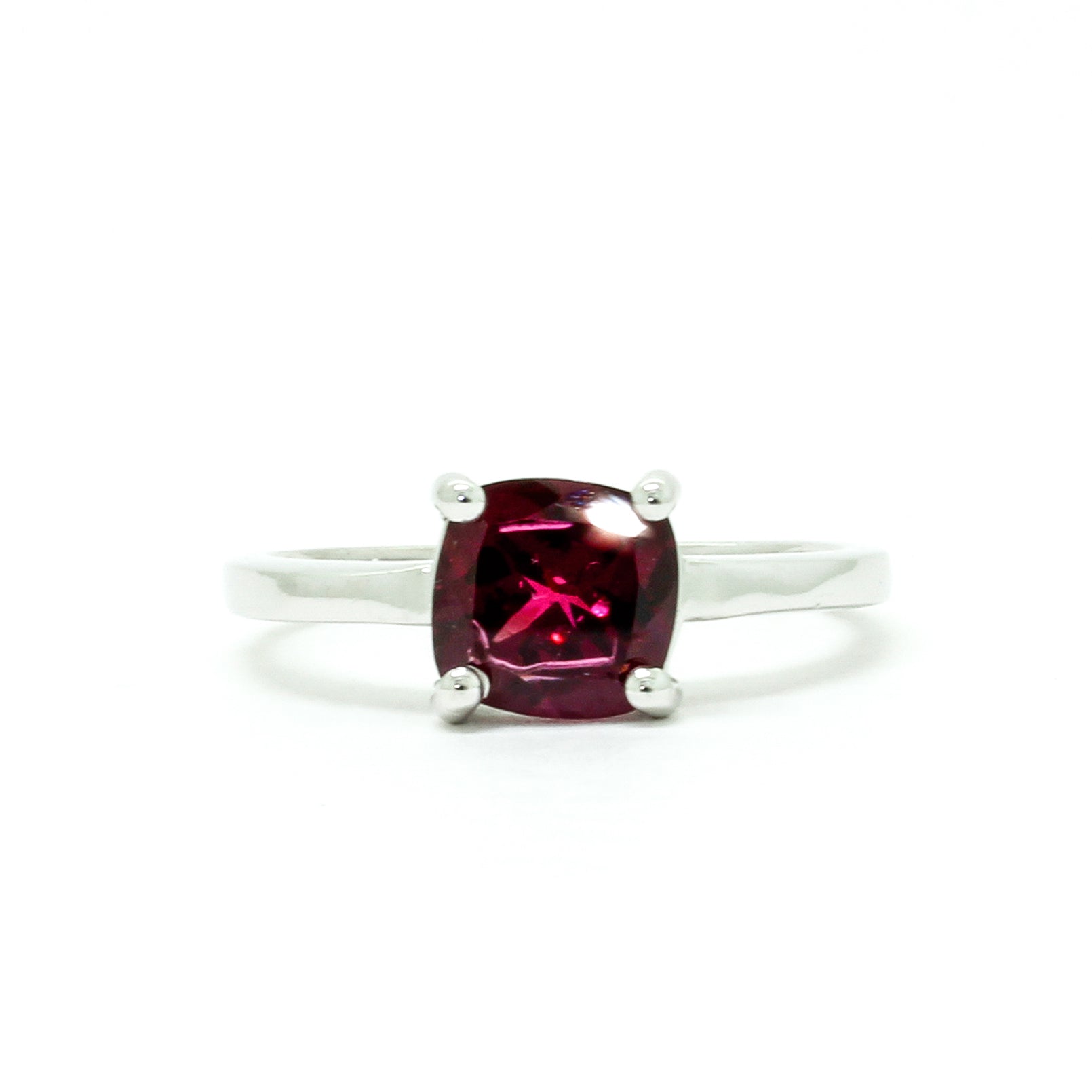 A product photo of a silver ring with a cushion-cut rhodalite centre stone sitting on a white background. The silver band is simple and smooth, connecting on either side of a square cushion-cut rhodalite stone held in place by four delicate silver claws. The rhodalite jewel is a deep velvet purple colour, reflecting a warm plum colours across its multi-faceted surface.