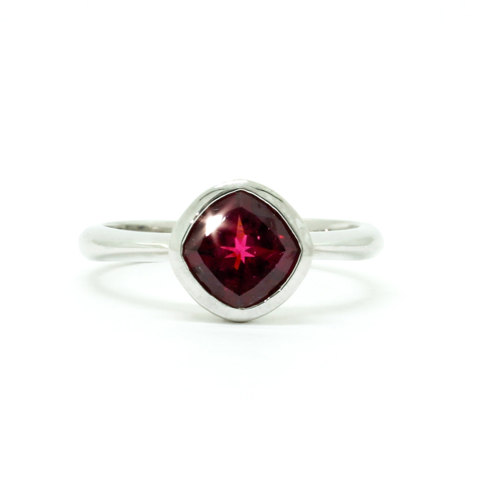 A product photo of a silver ring with a bezel-set rhodalite centre stone sitting on a white background. The silver band is simple and smooth, connecting on either side of a diagonally-oriented cushion-cut rhodalite stone surrounded by a solid frame of silver. The rhodalite jewel is a deep velvet purple colour, reflecting a warm plum colours across its multi-faceted surface.