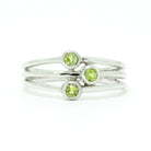 A product photo of a silver ring with 3 delicate bezel-set peridot stones sitting on a white background. The silver band is simple and smooth at the back before splitting into three prongs, each capped off with a delicate little peridot stone set within a silver frame. The prongs end at three different lengths, creating an interesting connecting line between the three stones. The peridot jewels are a shade of bright, pale green.