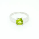 A product photo of a silver ring with a cushion-cut peridot centre stone sitting on a white background. The silver band is simple and smooth, connecting on either side of a square cushion-cut peridot stone held in place by four delicate silver claws. The peridot jewel is a shade of bright, vibrant green, reflecting chartreuse light across its multi-faceted surfaces.