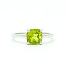 A product photo of a silver ring with a cushion-cut peridot centre stone sitting on a white background. The silver band is simple and smooth, connecting on either side of a square cushion-cut peridot stone held in place by four delicate silver claws. The peridot jewel is a shade of bright, vibrant green, reflecting chartreuse light across its multi-faceted surfaces.