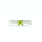 A product photo of a silver ring with a round-cut peridot centre stone sitting on a white background. The silver band is simple and smooth, connecting on either side of a small, roundl-cut peridot stone, held in place by four silver claws and a split band behind it. The peridot jewel is a shade of bright, vibrant green, reflecting chartreuse light across its multi-faceted surfaces.