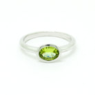 A product photo of a silver ring with a bezel-set peridot centre stone sitting on a white background. The silver band is simple and smooth, connecting on either side of a horizontally-oriented oval-cut peridot stone surrounded by a solid frame of silver. The peridot jewel is a shade of bright, vibrant green, reflecting chartreuse light across its multi-faceted surfaces.