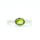 A product photo of a silver ring with a bezel-set peridot centre stone sitting on a white background. The silver band is simple and smooth, connecting on either side of a horizontally-oriented oval-cut peridot stone surrounded by a solid frame of silver. The peridot jewel is a shade of bright, vibrant green, reflecting chartreuse light across its multi-faceted surfaces.