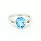 A product photo of a silver ring with a large bezel-set topaz centre stone sitting on a white background. The silver band is simple and smooth, connecting on either side of a vertically-oriented oval-cut topaz stone surrounded by a solid frame of silver. Before the band reaches the centre stone, it splits into two small horizontal silver oval frames on each side, each housnig a small diamond. The topaz jewel is almost the colour of a tropical sea, reflecting light across its multi-faceted surface.