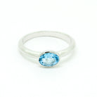 A product photo of a silver ring with a bezel-set topaz centre stone sitting on a white background. The silver band is simple and smooth, connecting on either side of a horizontally-oriented oval-cut topaz stone surrounded by a solid frame of silver. The topaz jewel is almost the colour of a tropical sea, reflecting bright blue light across its multi-faceted surface.