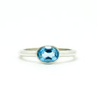 A product photo of a silver ring with a bezel-set topaz centre stone sitting on a white background. The silver band is simple and smooth, connecting on either side of a horizontally-oriented oval-cut topaz stone surrounded by a solid frame of silver. The topaz jewel is almost the colour of a tropical sea, reflecting bright blue light across its multi-faceted surface.