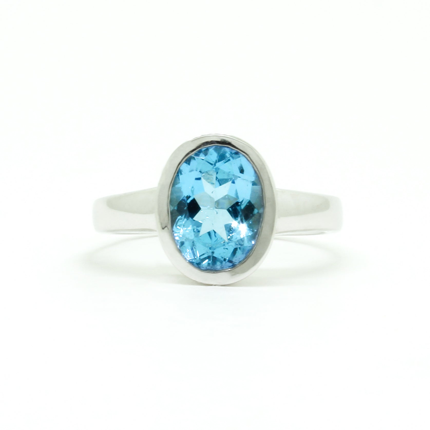 A product photo of a silver ring with a large bezel-set topaz centre stone sitting on a white background. The silver band is simple and smooth, connecting on either side of a vertically-oriented oval-cut topaz stone surrounded by a solid frame of silver. The topaz jewel is almost the colour of a tropical sea, reflecting bright blue light across its multi-faceted surface.