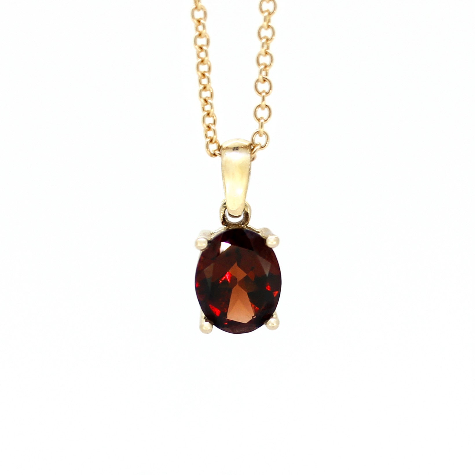 A product photo of a 9x7mm Oval Garnet Pendant in 9k Yellow Gold suspended against a white background. The impressively large and deeply-coloured oval-cut stone is contrasted by its overall minimalistic design, 4 simple golden claws holding the gem in place. It is suspended by a simple gold chain. The stone is a deep red, reflecting sanguine hues across its multi-faceted edges.