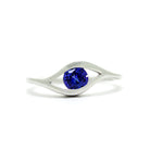 A product photo of a white gold tanzanite ring sitting on a white background. The band splits and curves around the single, round tanzanite centre stone, holding it in place like an eye. The tanzanite stone reflects violet blue and indigo colours from its many edges.