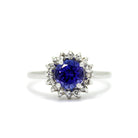 A product photo of a white gold tanzanite ring sitting on a white background. A flowery halo of diamond and white gold detailing frames the large, round centre stone. The deep blue tanzanite reflects violet blue and indigo colours from its many edges.