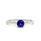 A product photo of a white gold tanzanite ring sitting on a white background. An otherwise simple white gold band is impressed upon by bubble-like detailing along its edges. The deep blue tanzanite stone reflects violet blue and indigo colours from its many facets.