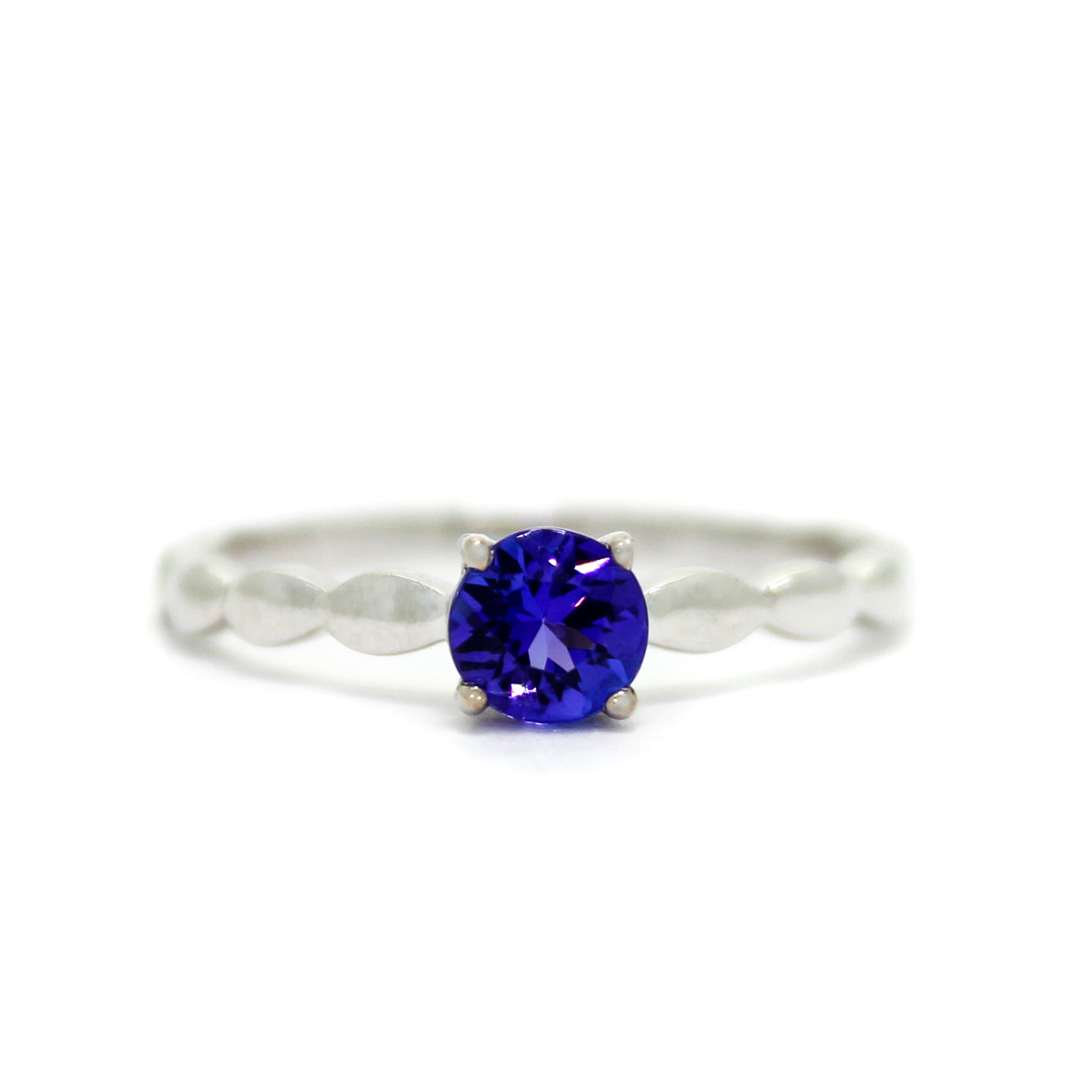 A product photo of a white gold tanzanite ring sitting on a white background. An otherwise simple white gold band is impressed upon by bubble-like detailing along its edges. The deep blue tanzanite stone reflects violet blue and indigo colours from its many facets.