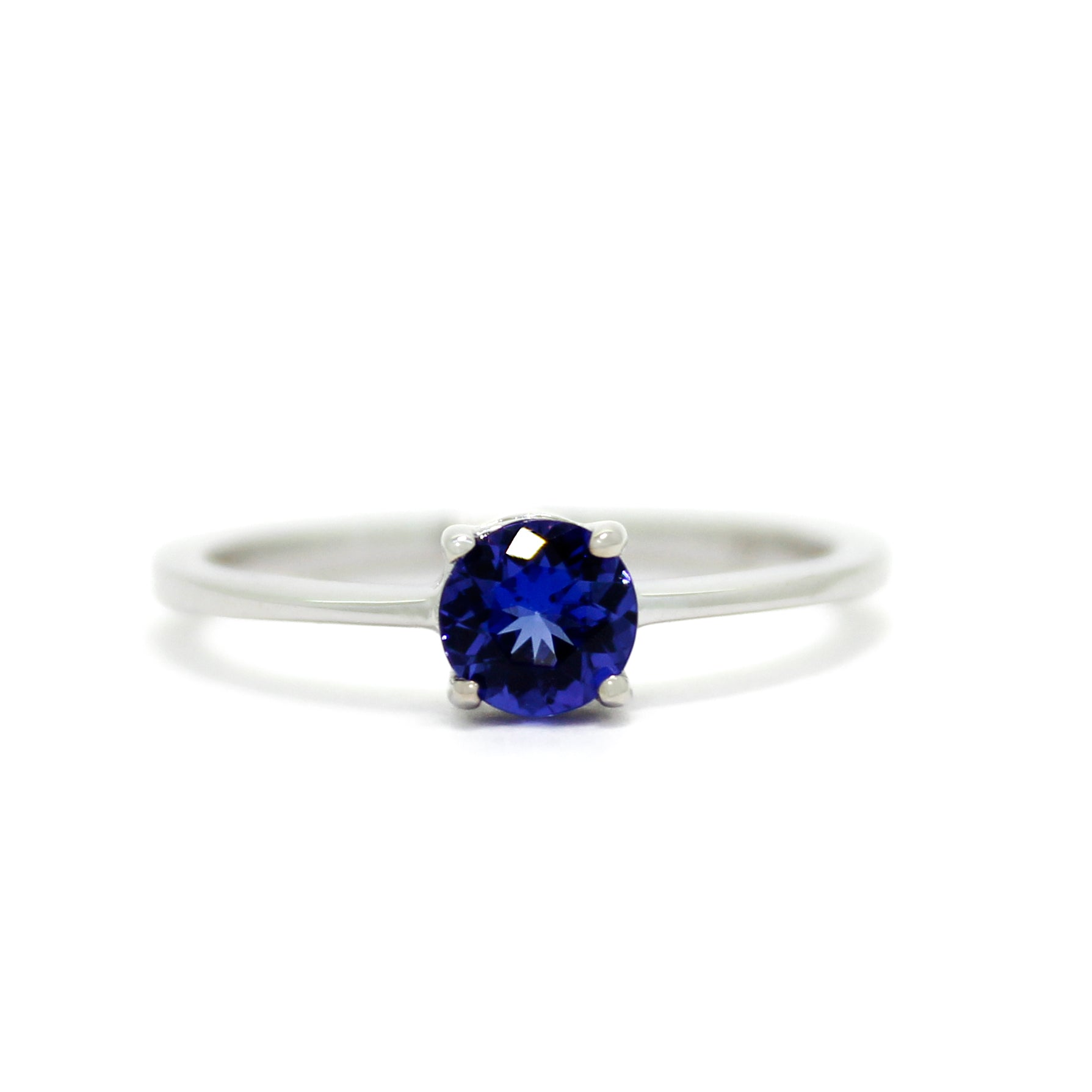 A product photo of a white gold tanzanite solitaire ring sitting on a white background. The band is smooth and simple, with the centre stone held in place by 4 small claws. The deep blue tanzanite stone reflects violet blue and indigo colours from its many edges.