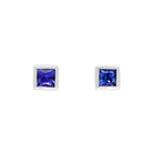 A product photo of white gold tanzanite stud earrings sitting on a white background. The square stones are framed by a thick layer of white gold. The deep blue tanzanites reflect violet blue and indigo light from their many edges.