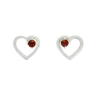 A product photo of two silver heart-shaped stud earrings sitting on a white background. A delicate, 2.5mm round red garnet is nestled in the top right corner of each heart-shaped frame.