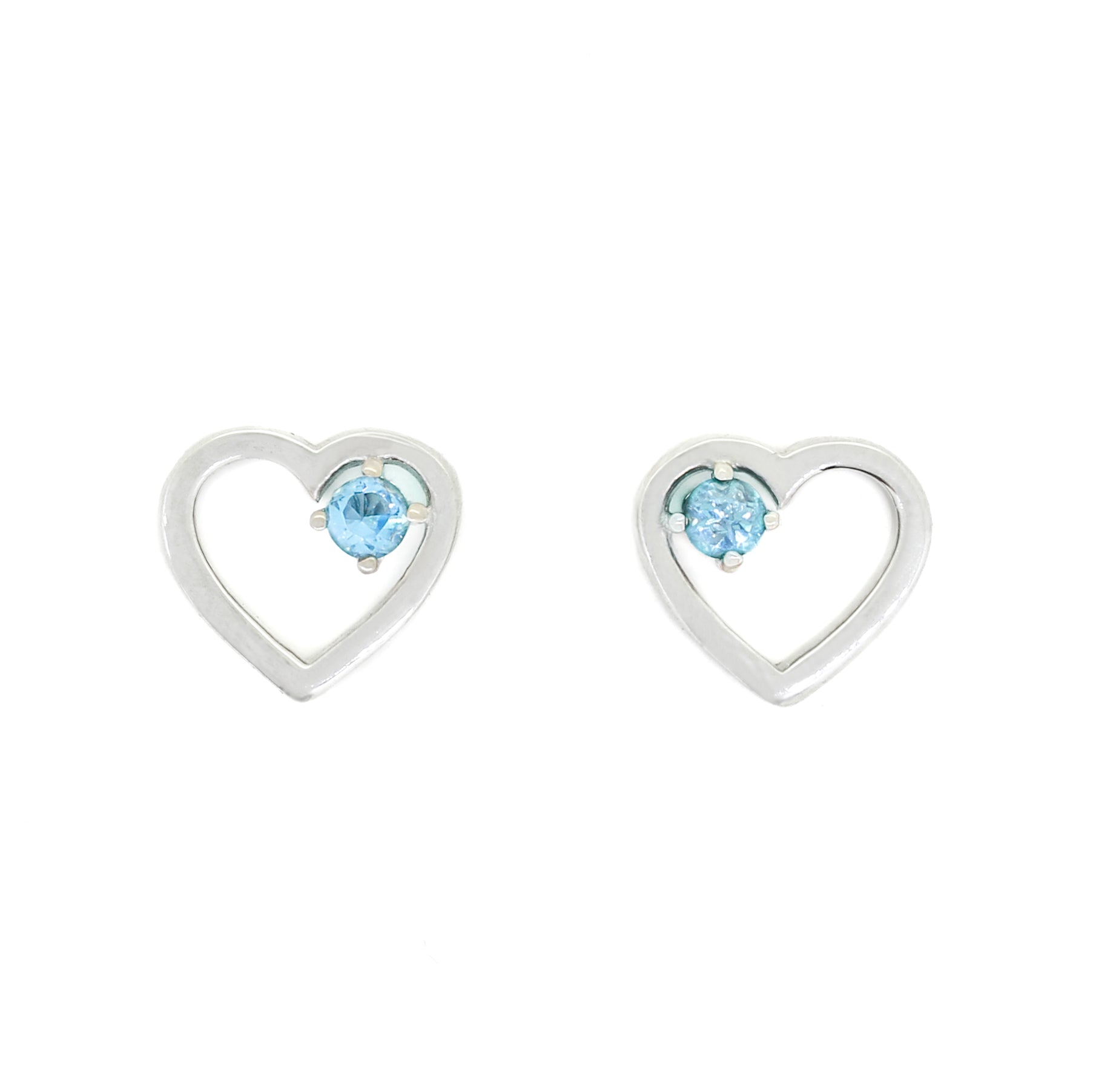 A product photo of two silver heart-shaped stud earrings sitting on a white background. A delicate, 2.5mm blue topaz stone is nestled in the top right corner of each heart-shaped frame.