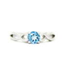 A product photo of an ornate silver ring with a blue topaz centre stone sitting on a white background. The silver band splits halfway along its length, becoming twisting and serpentine in appearance before meeting on either side of the dazzling blue 5mm stone, which is held in place by 4 silver claws.