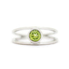 A product photo of a split-band silver ring with a bezel-set peridot centre stone sitting on a white background. The silver band splits at the base of the ring, separating to meet at the top and bottom of the light green round-cut centre stone, framed in a thick layer of silver, holding it in place.