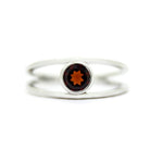 A product photo of a split-band silver ring with a bezel-set garnet centre stone sitting on a white background. The silver band splits at the base of the ring, separating to meet at the top and bottom of the deep red round-cut centre stone, framed in a thick layer of silver, holding it in place.