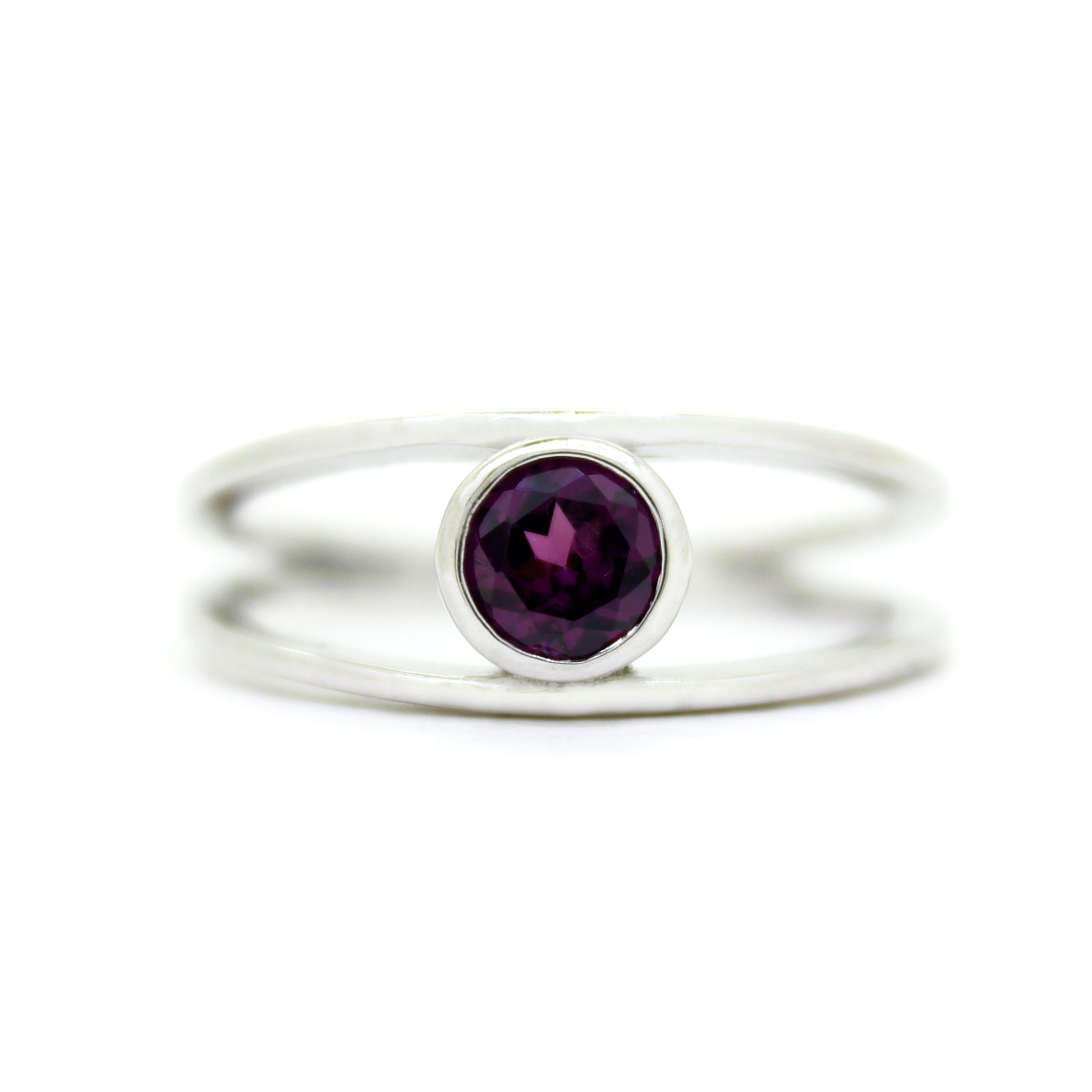 A product photo of a split-band silver ring with a bezel-set rhodalite centre stone sitting on a white background. The silver band splits at the base of the ring, separating to meet at the top and bottom of the plum-coloured round-cut centre stone, framed in a thick layer of silver, holding it in place.