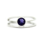 A product photo of a split-band silver ring with a bezel-set iolite centre stone sitting on a white background. The silver band splits at the base of the ring, separating to meet at the top and bottom of the deep, dark blue round-cut centre stone, framed in a thick layer of silver, holding it in place.