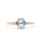 A product photo of a rose gold aquamarine ring sitting against a white background. The rose gold band is plain and smooth, and the centre oval-cut light blue aquamarine stone is framed by a single white diamond on either side.