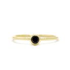 A product photo of a delicate yellow gold stacking ring with a tiny, bezel-set black diamond in the centre sitting on a white background. The band is slim and thread-like, with the focus drawn to the petite 3mm glinting black centre stone.