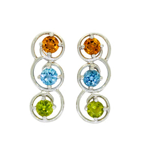 A product photo of two multi-gemstone earrings in 9k white gold on a white background. The earrings are made up of 3, wave-like loops of white gold each, with each loop adorned with a round-cut colour gemstone. From the top to bottom of each earrings are orange citrines, blue topaz and warm green peridots.