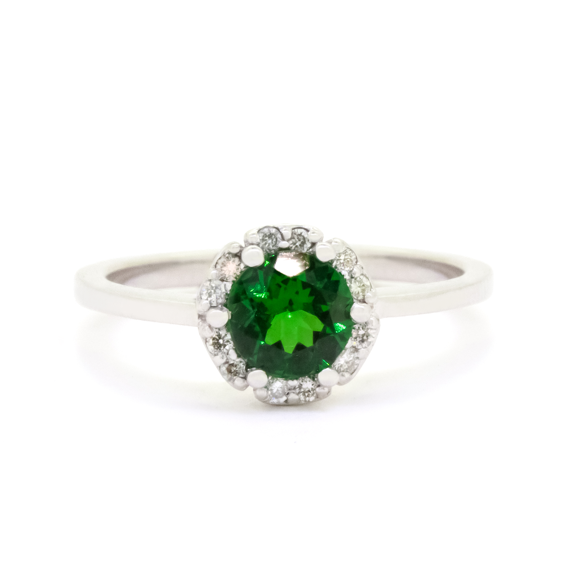 A product photo of a classic diamond halo tsavorite ring in solid 9k white gold on a white background. The rounded band is smooth and minimalistic. The deep, verdant-green tsavorite centre stone is held in place by 6 claws and reflects light off of its many facets. The green gemstone colour be a good emerald substitute.