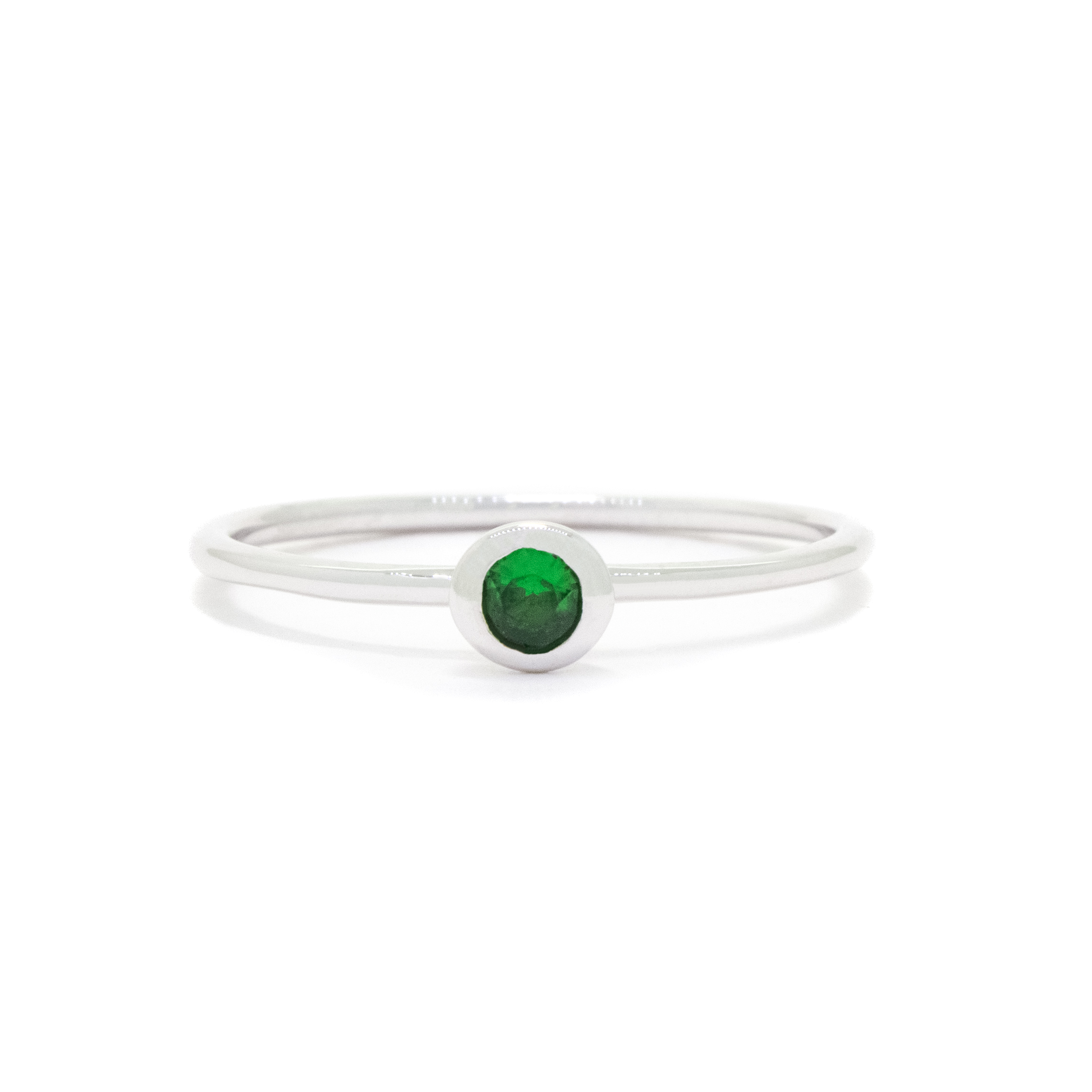 A product photo of a delicate white gold stacking ring with a tiny, bezel-set tsavorite in the centre sitting on a white background. The band is slim and thread-like, with the focus drawn to the petite 3mm glinting green centre stone. The green gemstone colour be a good emerald substitute.