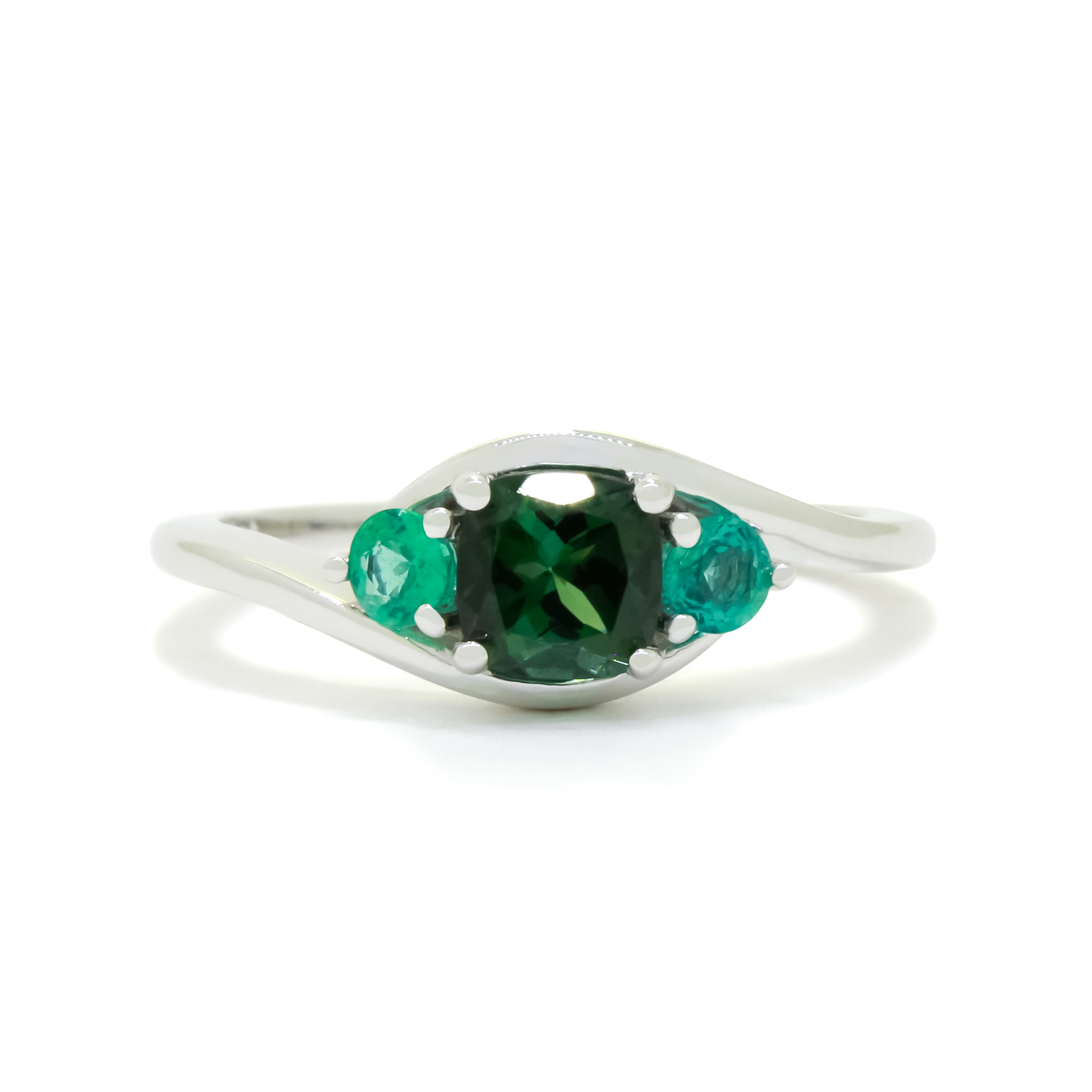 A product photo of a mystical tourmaline and emerald fantasy ring in solid 9k white gold on a white background. The deep, forest-green tourmaline is held in place by 4 claws and reflects light off of its many facets, while the emeralds on either side are held by 3 each. The smooth white gold band curves elegantly, meeting at the top and bottom of the tourmaline and emerald arrangement. The green gemstone colour be a good emerald substitute.