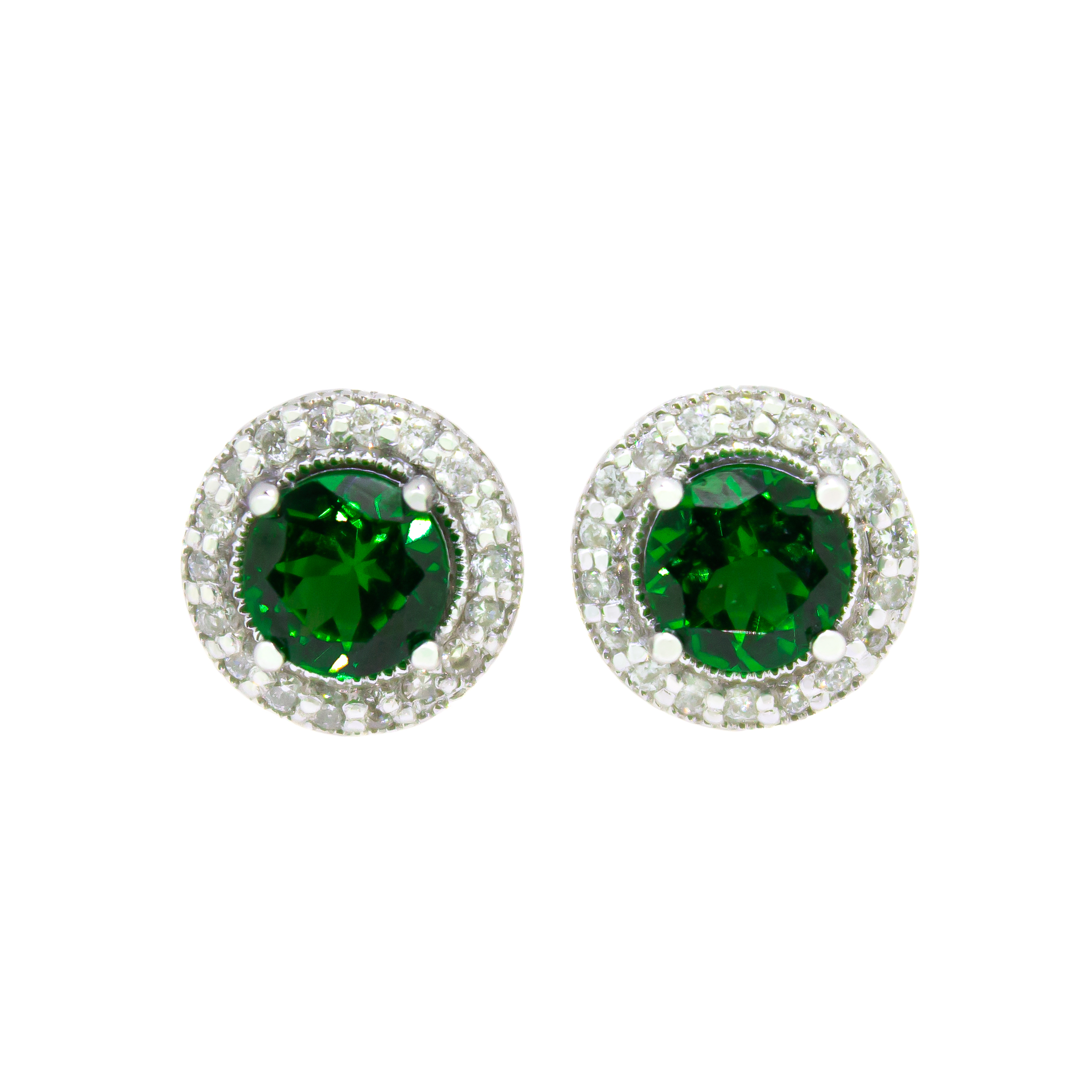 A product photo of a pair of white gold tsavorite earrings sitting against a white background. The pair of circle-cut green tsavorite are surrounded by thick frames of ornately detailed white gold and diamond detailing. The green gemstone colours make these earrings a good alternative or substitute for emerald.