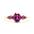 A product photo of a dainty and elegant yellow gold ring with a stunning trio of grape garnet and amethysts sitting against a white background. The largest of the stones sits in the centre, a 7x5mm oval grape garnet - hugged by two smaller pear-shaped amethyst stones on either side.