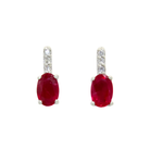 A product photo of a pair of white gold ruby earrings sitting against a white background. The simple oval-cut stones are contrasted by the white diamond trio protruding in a straight line above each burgundy magenta stone, encased in white gold.