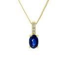 A product photo of a yellow gold sapphire pendant suspended against a white background. The simple oval-cut stone is contrasted by the white diamond trio protruding in a straight line above the magenta ruby, encased in yellow gold.