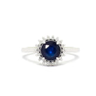 A product photo of a white gold sapphire ring sitting on a white background. A flowery halo of diamond and white gold detailing frames the large, round centre stone. The deep blue sapphire reflects blue and azure colours from its many edges.