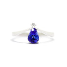 A product photo of a delicate tanzanite teardrop ring with a single diamond accent in 9ct white gold sitting on a white background. The smooth, slim white gold band curves elegantly to meet a tiny, 0.015ct white diamond jewel, which sits atop a 7x5mm pear shaped indigo blue tanzanite gemstone.