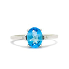 A product photo of a white gold blue topaz and diamond ring sitting against a white background. The white gold band is plain and smooth, and the centre oval-cut light blue topaz stone is framed by a single white diamond on either side.