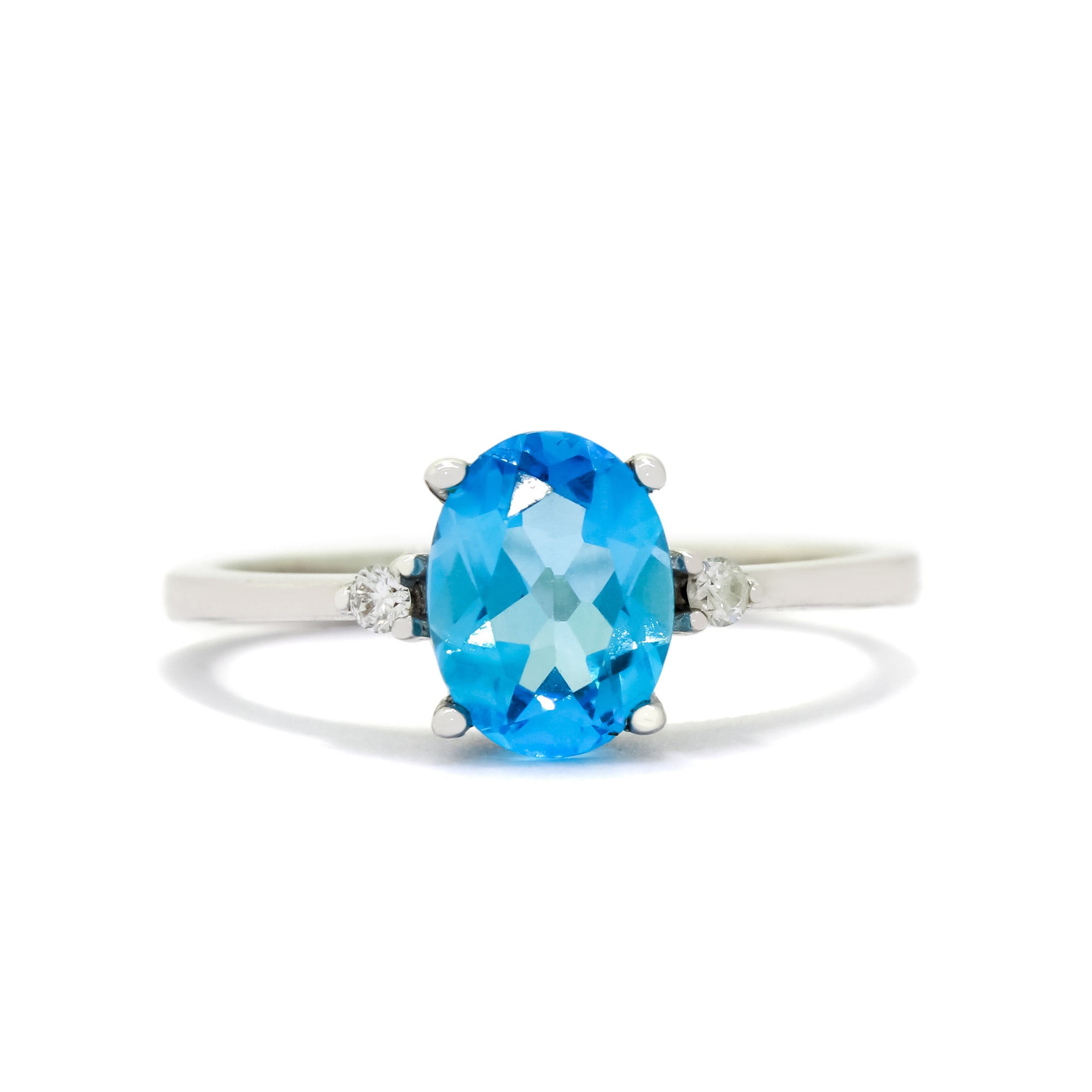 A product photo of a white gold blue topaz and diamond ring sitting against a white background. The white gold band is plain and smooth, and the centre oval-cut light blue topaz stone is framed by a single white diamond on either side.