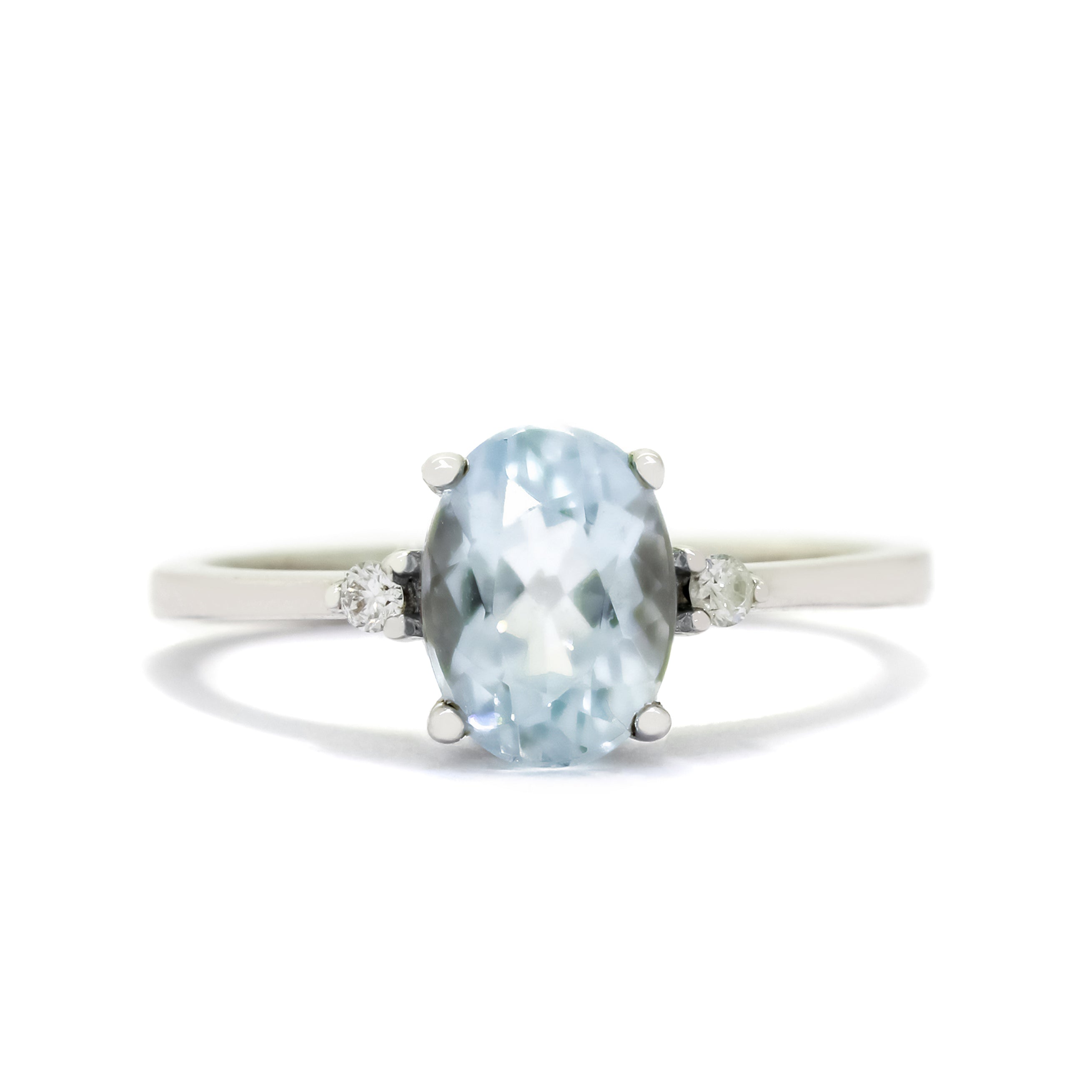 A product photo of a white gold blue aquamarine and diamond ring sitting against a white background. The white gold band is plain and smooth, and the centre oval-cut light blue aquamarine stone is framed by a single white diamond on either side.