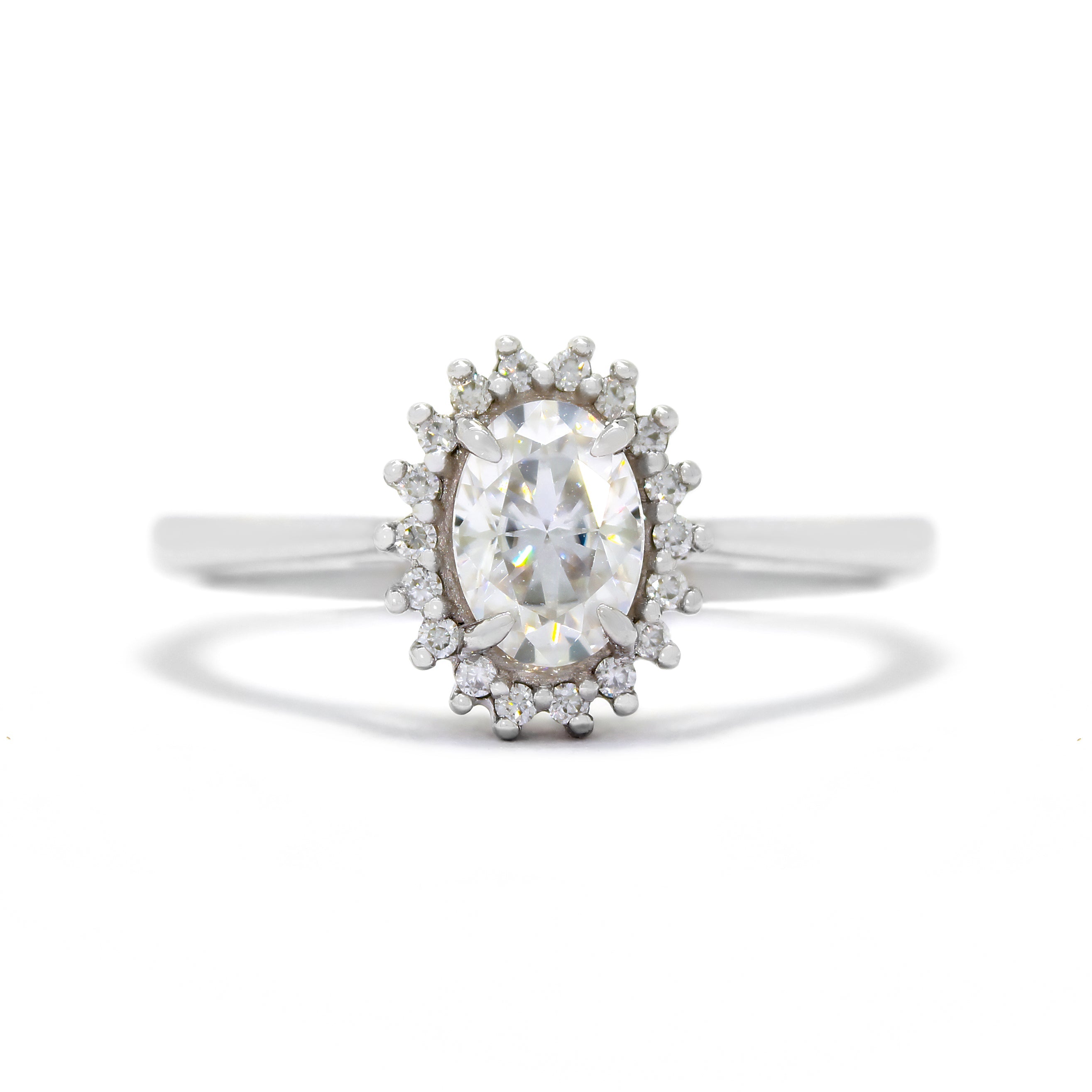 A product photo of a moissanite halo ring in 9ct white gold on a white background. A large, dazzling oval moissanite stone in the centre reflects rainbow-coloured light, and is framed by a ring of smaller white moissanites. The band is simple and rounded.