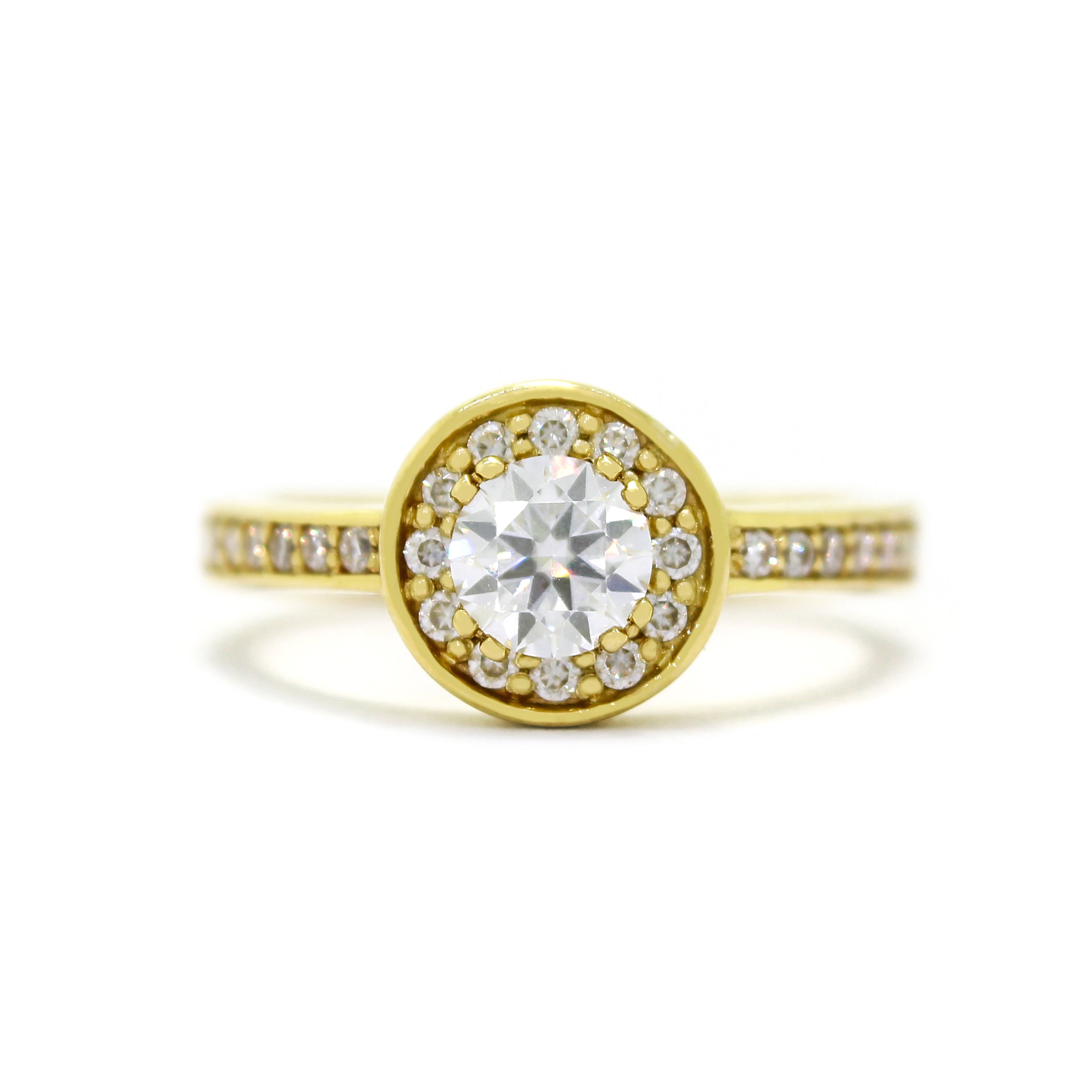 A product photo of a classic moissanite halo ring in solid 9ct yellow gold on a white background. The moissanite halo is framed by a thick band of solid 9k yellow gold, and the rounded band is smooth and minimalistic. The dazzling white moissanite centre stone is held in place by 4 claws and reflects light off of its many facets.