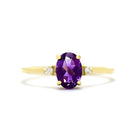 A product photo of a yellow gold amethyst ring sitting against a white background. The yellow gold band is plain and smooth, and the centre oval-cut deep purple amethyst stone is framed by a single white diamond on either side.